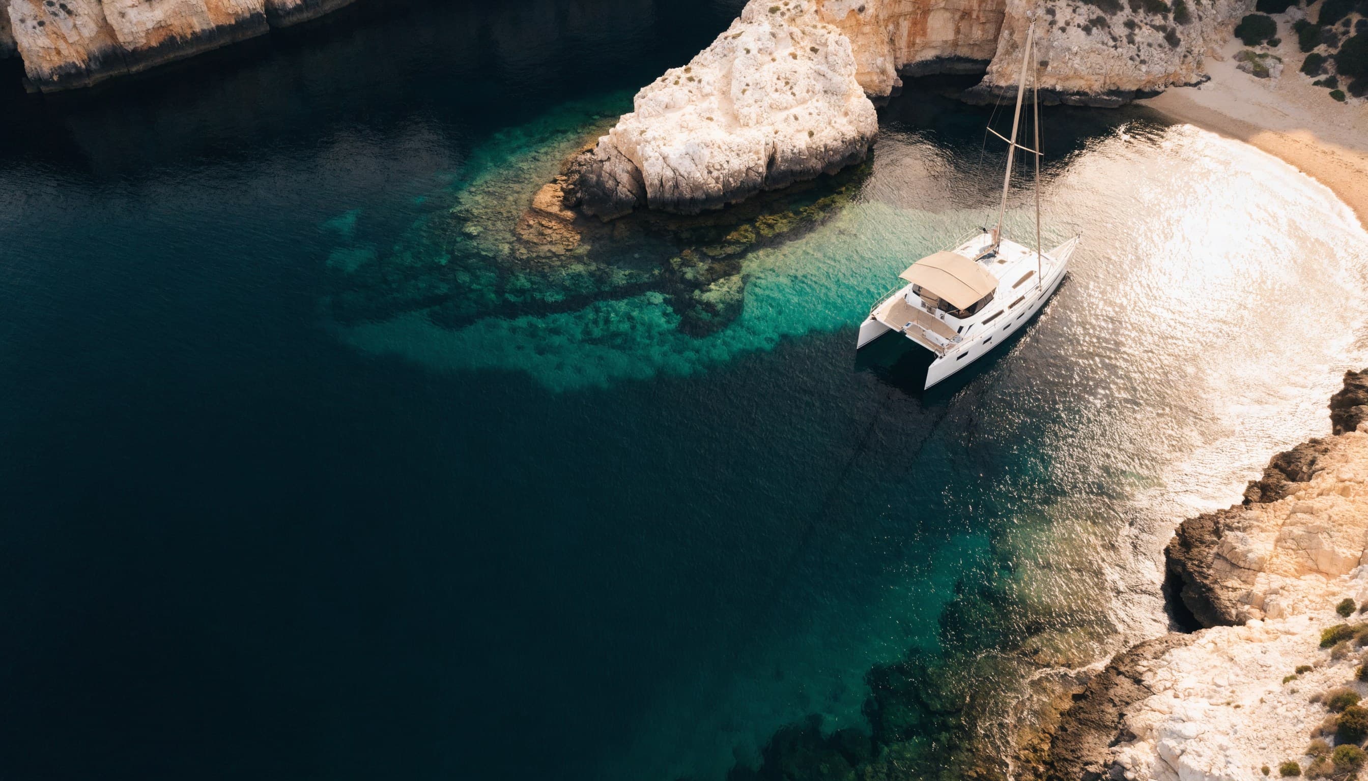 Aerial view of a luxury catamaran anchored in a dramatic turquoise Mediterranean cove